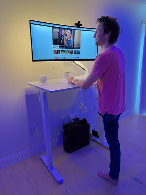 Josh is standing at an electric desk looking at a horizontal computer monitor where the TV series "Best Foot Forward" on Apple TV  is featured in the background. Josh is wearing a t-shirt and jeans. 