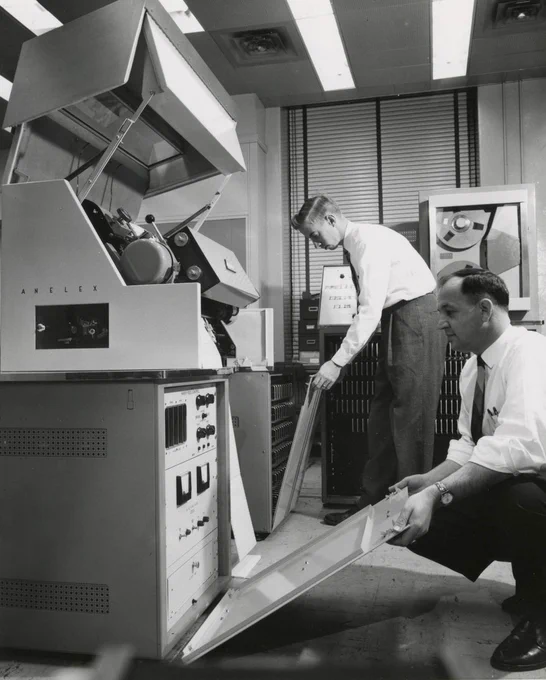 CBI Image of the Day. Black and white portrait rectangular orientation Annelex made pioneering high-speed printers, depicted here are two men looking at an Annelex with the hood up and panels removed exposing dials and controls. A tape drive is in the background. Florescent lights overhead.
