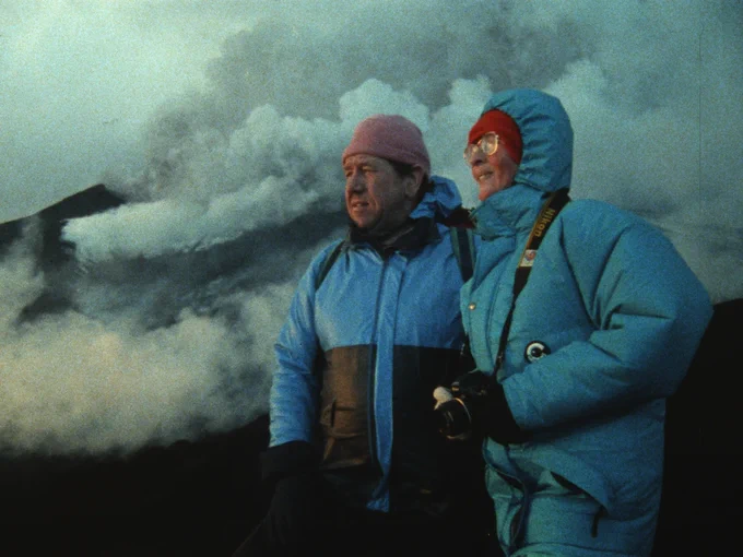 A man and a woman, wearing thick insulated blue coats, gloves and colourful hats stand before a smoking volcano. Their faces are illuminated. 