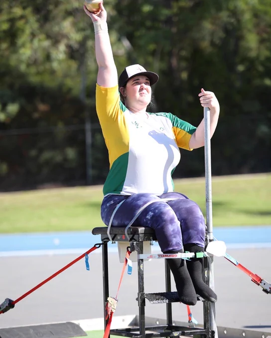 Woman seated in wheelchair throwing shotput.