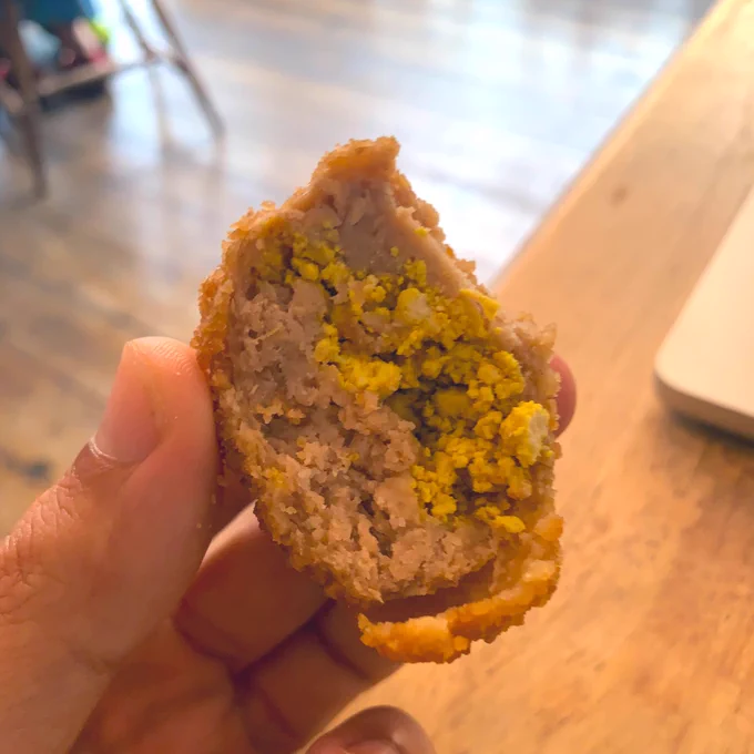 My hand holding half of a little vegan scotch egg. It shows the inside, which looks like sausage and boiled egg yolk – but it's tofu