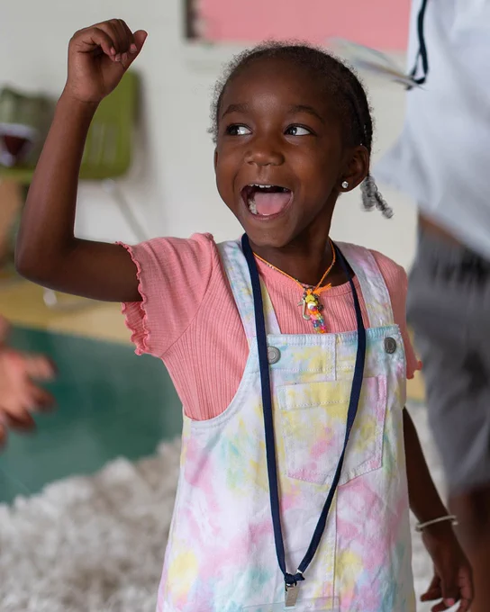An excited young child in tie-dye overalls cheers. Their hand is over their head, and they are smiling widely. 