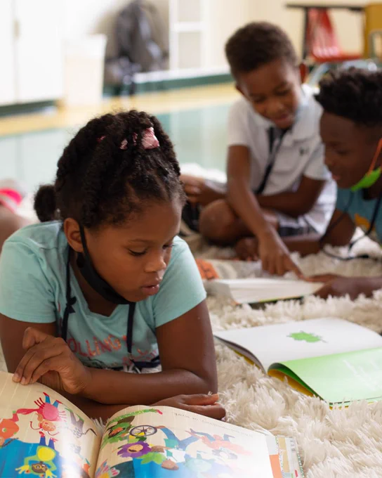 Three children are reading books on a carpet. 