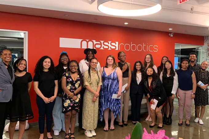 A group of people of varying ages, genders, and races stand in front of a red wall with the MassRobotics logo. 