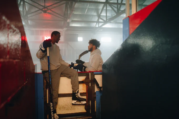 A hockey player sits on an arena board wearing skates and holding a hockey stick. He talks to another player who is standing on the ice in the film Black Ice.
