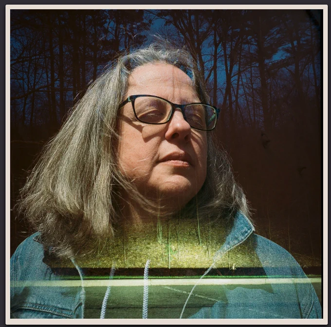 A double exposure of a portrait of Kate Price and the wooded area near a highway rest stop. Kate's gray hair frames her face, where her eyes are closed as her head turns to the left. She's wearing a blue jean jacket. The trees of the wooded area are silhouettes against a dark blue, dusk sky. The pavement and leaf-covered ground are illuminated by car headlights.
