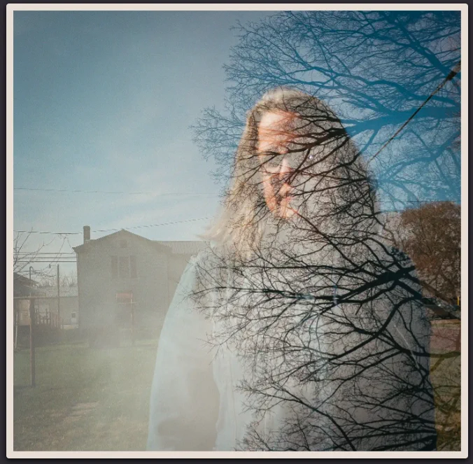 A double exposure of Kate overlaid with the woods at the rest area. Kate stands in front of her childhood home, which is seen over her right shoulder. She wears a blue jean jacket. Entering the frame on the right are the leafless branches of trees that reach out and partially obscure Kate.