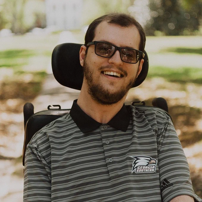 Headshot of Joshua. A young man  with brown hair, sunglasses, a beard wearing a striped collar shirt. He is sitting in a wheelchair outside.