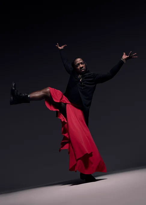 A person wearing a black top stands on one leg with their hands in the air. They have a red, flowy skirt on and are wearing black boots. 
