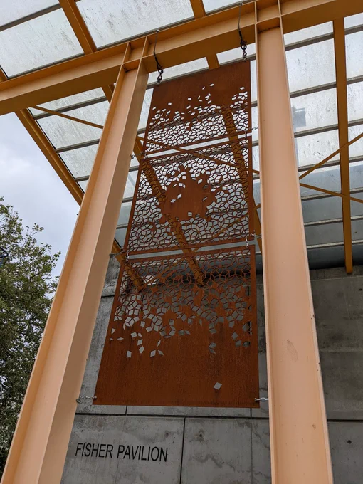 Three rusty 1/4" panels hanging between two yellow I-beam columns. They each have a Penrose tiling lattice cut out of them, with randomized tiles left solid. On the top and bottom panel, these form a linear gradient, while the middle panel is radial.
