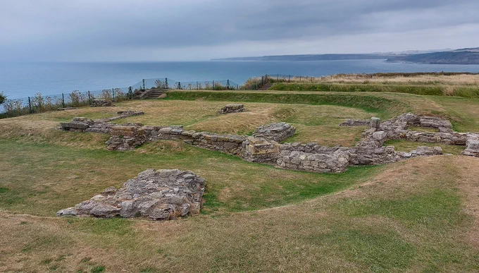 A colour landscape photograph showing the remains of a roughly square enclosure on a clifftop, with the sea behind. Its perimeter is marked with an earthen bank, and the low remains of stone walls and structures stand inside. The grass is parched and the view dramatic.
