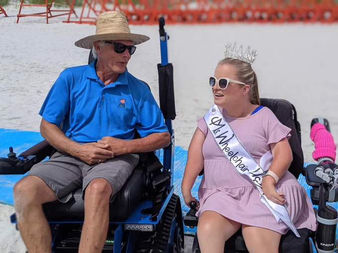 Sara in her crown and sash speaks to a man wearing a beach hat and sunglasses who is using n accessible power beach chair.
