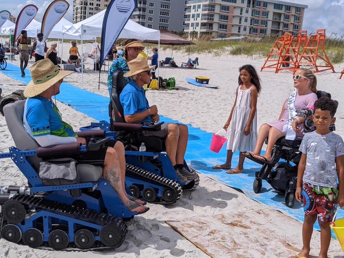 Sara talks to two people who are sitting in adapted power beach chairs, while in her standard power chair on a beach mat.