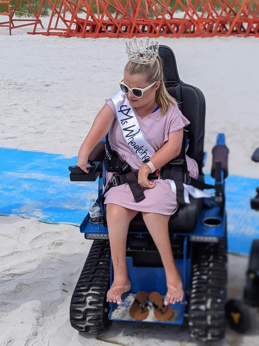 Sara in her crown and sash and sunglasses uses the accessible power beach wheelchair. Her flip flops are off and her feet are free.