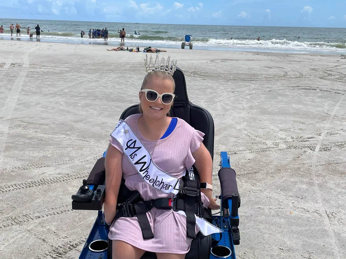 Sara smiles in her Ms. Wheelchair USA crown and sash and sunglasses while she poses in front of the ocean in an accessible beach power wheelchair.