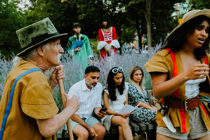 Members of Dauntless City Theatre perform outside, while three members of the audience sit on a bench between them. Two actors are in the foreground, and two actors are in the background, and there is a party of three seated between them in the midground. There is a bank of wild purple sage behind the people sitting on the bench. 

