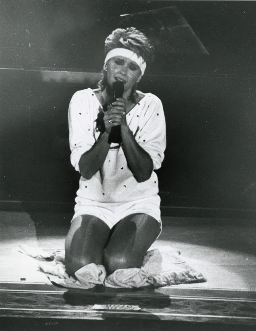 Olivia Newton-John kneels on stage while singing during her Physical Live concert tour in 1982. She's wearing a 80s workout outfit with a headband. The photo is in black and white. 