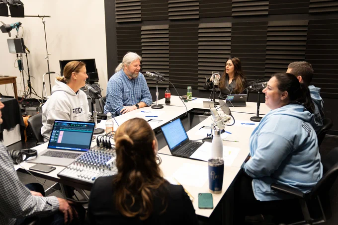 Seven people sit around a table in a podcast studio.