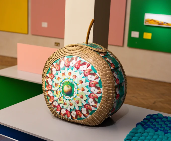 Apple Laundry Basket by Lois Wapole. Made in London in 1995. Description: Basket made of coiled willow and string with apple juice cartons as a central theme and structure on the front and back, with a lid with a wooden stick to act as the handle.