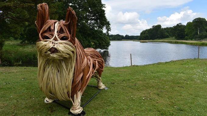 Photo of wicker corgi, standing on grass in front of lake.