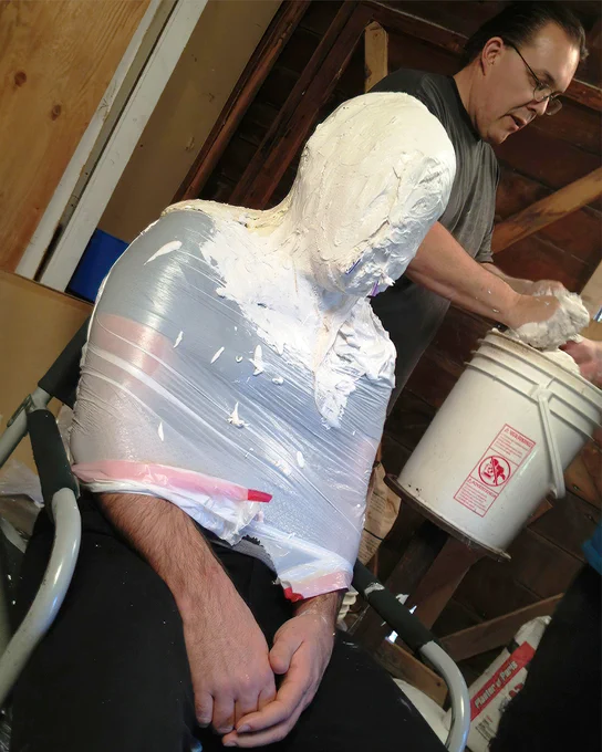 Man's head is covered in plaster with two straws sticking out if his nose area. Odin mixes plaster in the background.