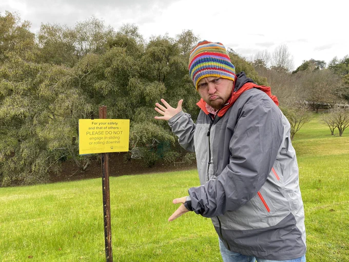 Person grumpily gesturing to a sign that reads For your safety and that of other PLEASE DO NOT engage in sliding or rolling downhill