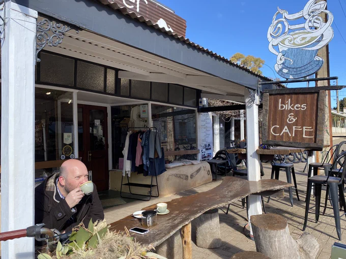 Person sipping a cup of tea in the sunshine outside a shop with a sign saying Bikes & Cafe