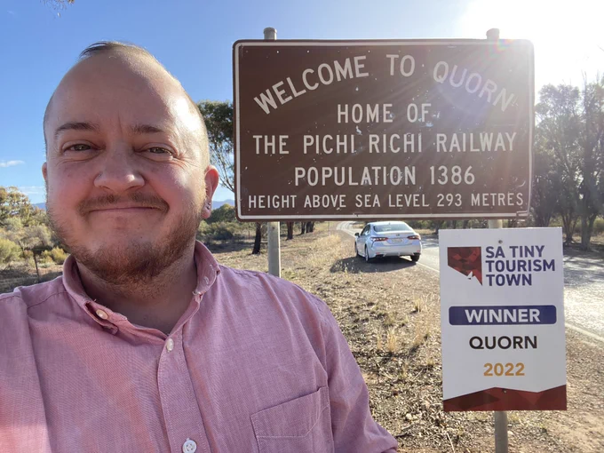 Selfie of smiling person beside a sign reading Welcome to Quorn home of the Pichi Richi Railway, population 1386, height above sea level 293 metres