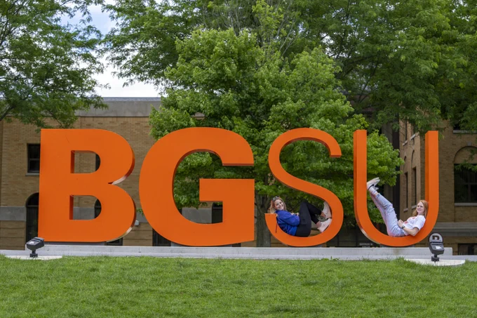 two bgsu students sit in the "S" "U" portion of the orange BGSU letter statues on campus in the summer