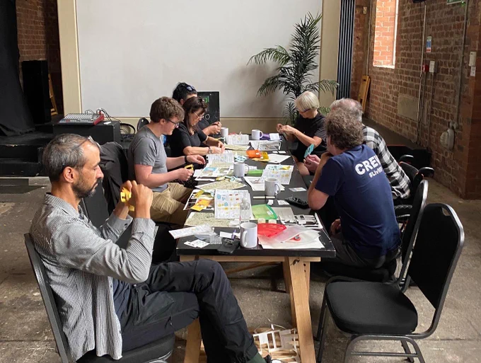 Workshop participants sit together around a long table. The group are all looking away from the camera concentrating on making foldable paper microscopes to view the bioplastics. The table is very full of paper and card parts of the microscopes, as these are in various stages of assembly, and there are also mugs of hot drinks on the table.