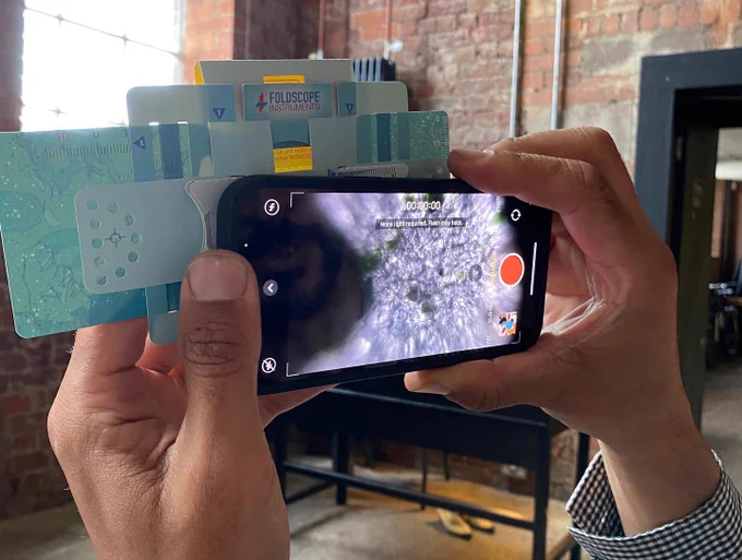 A participant is using their Foldscope paper microscope, holding this up to the light of the station window,  to view some algae bioplastic. The microscope is attached to a mobile phone and the microscopic image of the material is shown on the phone’s screen with a purple, grey and white marbled image flecked with abstract green shapes.
