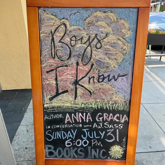 A chalk art sidewalk sign advertising an author event is outside Books Inc in San Francisco. The sign has a warm wood colored frame. The artwork depicts pastel colored clouds and the features book’s title in negative space. Text reads Boys I Know, author Anna Gracia in conversation with A. J. Sass, Sunday July 31 6:00pm Books Inc.