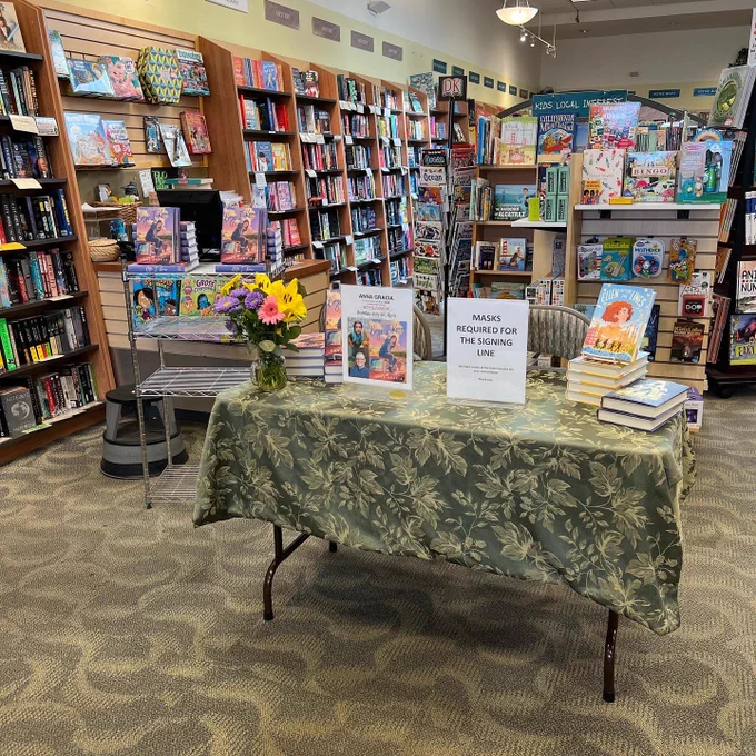 The view of an event table inside Books Inc bookstore in San Francisco. The table has flowers on it, plus copies of the features book, Boys I Know by Anna Gracia. There are also book copies of Ellen Outside the Lines and Ana on the Edge by A. J. Sass, the event host. The table has an event poster and also a sign indicating that masks must be worn when approaching the signing table. Many full shelves of books are in the background.