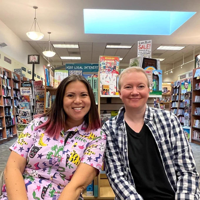 Biracial author Anna Gracia smiles while sitting next to white nonbinary author A. J. Sass. Anna wears a pink jumpsuit with lots of cartoon prints while A. J. wears a blue and white checkered button-up with a black tshirt underneath. There are bookshelves and displays behind them. The location is Books, Inc. in San Francisco.