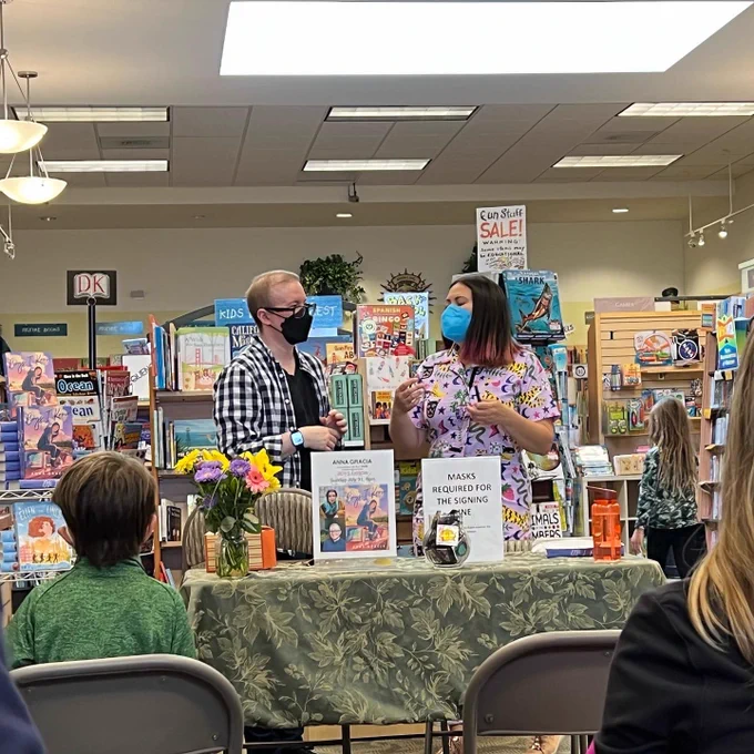 Biracial author Anna Gracia chats with white nonbinary author A. J. Sass. Anna wears a pink jumpsuit with lots of cartoon prints and a blue face mask while A. J. wears a blue and white checkered button-up with a black tshirt underneath and a black face mask. There are bookshelves and displays behind them and a table displaying a poster of their upcoming book event, plus the backs of some audience members, in front of them. The location is Books, Inc. in San Francisco.