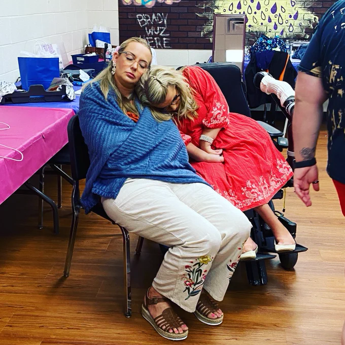 Sara wearing a red dress is leaning on Angelina who has her head on Sara’s. Angelina is wrapped up in a blue blanket.