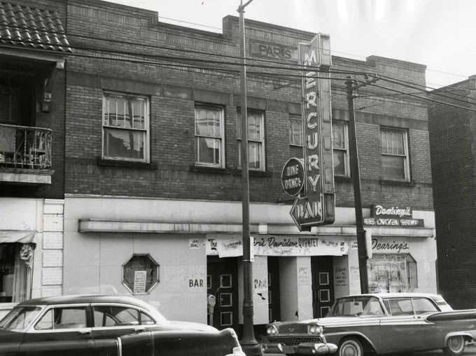 1940s-50s cars parked in front of an old commercial building with a blade sign reading "Mercury Bar Dine Dance" and a smaller sign reading "Dearing's Ribs Chicken Shrimp." An iron balcony to the left is sheltered by a tile-roofed awning supported by brackets. The building itself has the name "Paris" carved in a stone nameplate set in the brick facade just below its roofline.