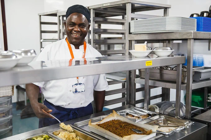 Sal, wearing chef whites, navy blue apron and hat, and an orange St Catherine's Hospice lanyard, smiling at the camera whilst stood in the kitchen, serving food.
