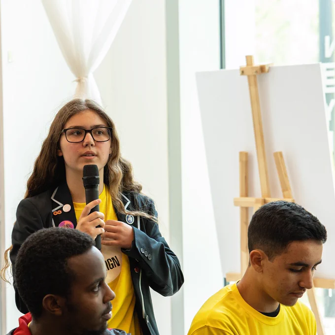 One Young Person speaking on microphone in yellow Fair Energy shirt. Two Young People sitting looking around wearing Yellow Fair Energy shirt.