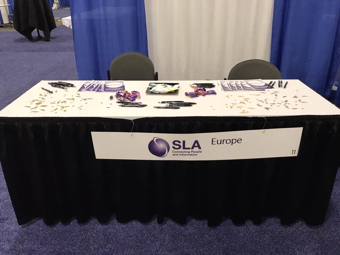 Table covered in a black cloth with a sign reading SLA Europe. Table top has merchandise like pens and chocolates.