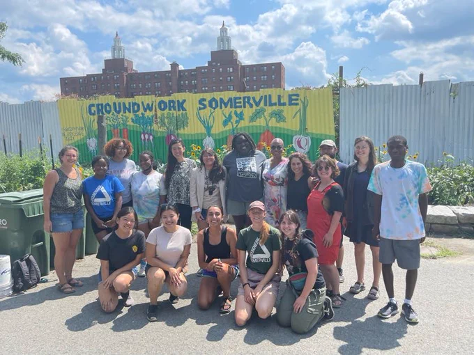 Congresswoman Pressley poses for a photo with staff and volunteers at Groundwork Somerville. There is a yellow and green mural behind them that reads "GROUNDWORK SOMERVILLE."