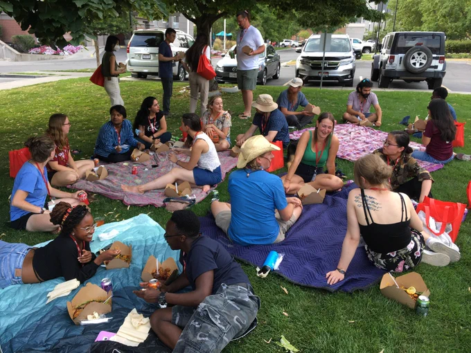 A group of graduate students and instructors sitting on blankets on the grass, having a picnic, talking, and laughing.