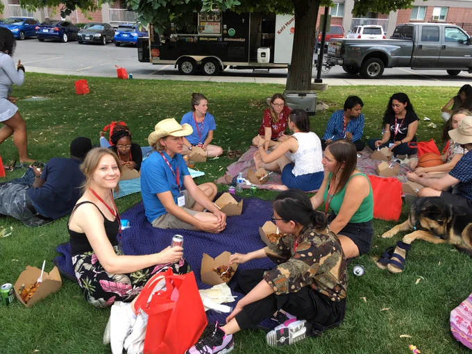 A group of graduate students and instructors sitting on blankets on the grass, having a picnic, talking, and laughing.