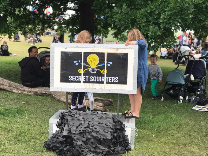 The image shows families sitting in a park with two girls standing behind a display case saying "Secret Squirters". Image by The Bureau of Silly Ideas.