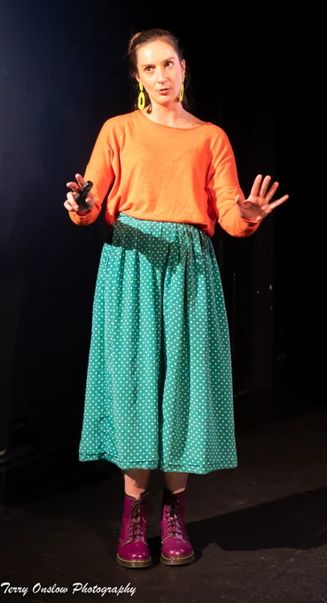 A woman in an orange top, green skirt, and purple boots is talking to the audience.
