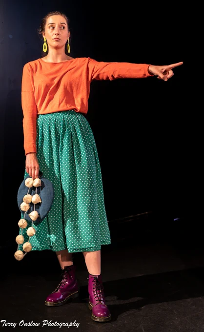 A woman in an orange top, green skirt and purple boots is holding a beret and a string of garlic while pointing at something.