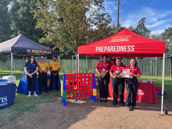 UC Merced Police Department and Emergency Management staff standing underneath their booth tents outdoors and smiling at the camera.