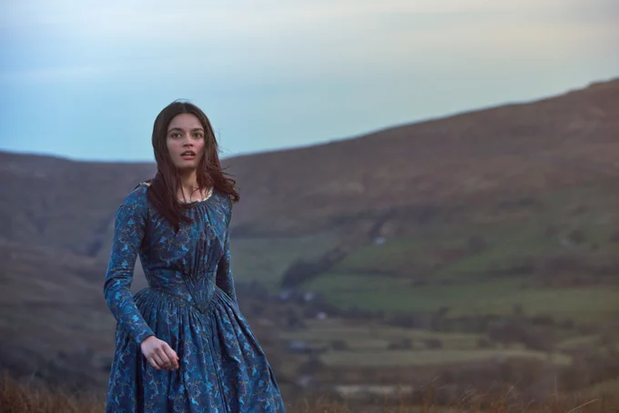 Emma Mackey wears a blue Victorian gown in the film Emily. She stands in a field with green hills, blue sky and her hair blowing in the wind.