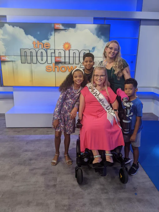 Sara Gaver, wearing her Ms. Wheelchair USA crown and sash, with her best friend Angelina and Angelina’s 3 kids in front of the Morning Show sign.