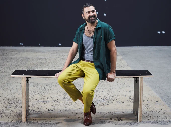  Jeff Khan sits on a bench and smiles. He wears a grey singlet with a green unbuttoned shirt over the top shirt and yellow jeans.